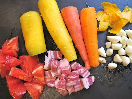 Dette trenger du til gryta: 1-2-3 kjøttfulle tomater (en stor eller 3 mindre), 4 store gulrøtter (jeg har gulrøtter i forskjellige farger fra Korsvold gård, men det trenger du altså ikke. Velg gulrøtter med mye smak og skjær dem i tykke stykker slik at de ikke koker ut. De skal beholde formen i den lange kokeprosessen. Alternativt kan du tilsette halvparten fra start og resten når det gjenstår en time. Da vil du ha noen gulrøtter som er mindre kokte. Revet skall av 1/4 appelsin (ikke få med det hvite. En potetskreller er perfekt til dette formålet), 2 hele hvitløk, skrelt. Så trenger du 200 g godt bacon i tykke stykker. Velg kvalitetsbacon fra Idsøe eller Haugen Gardsmat hvis du kommer over det. 